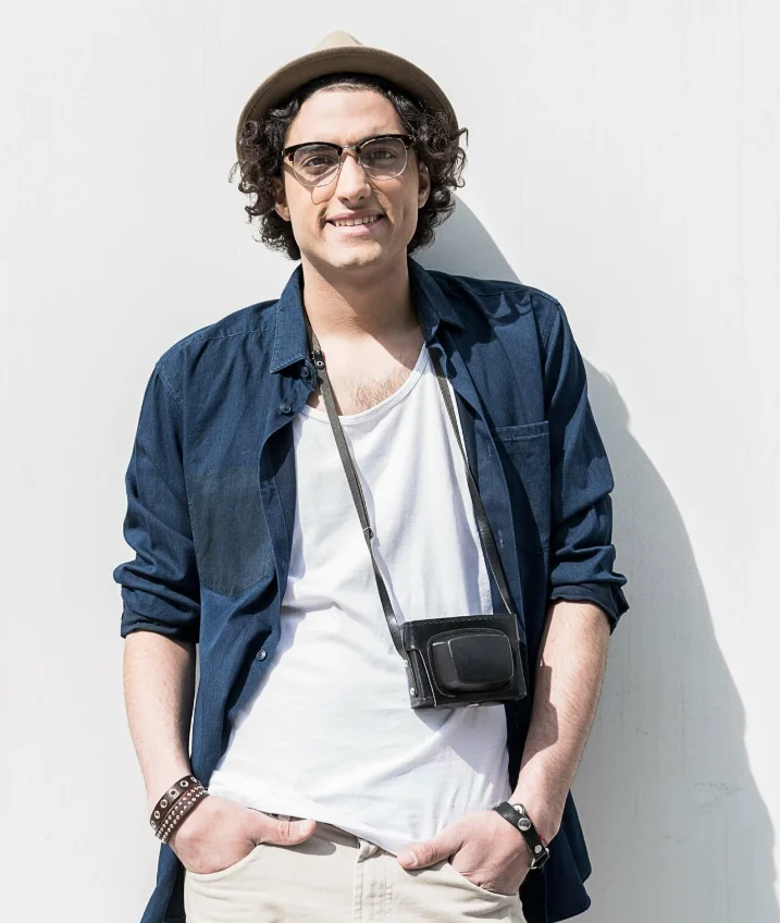 Positive vibes. Portrait of cheerful young man is leaning on white wall. His hands are in pockets of his pants. He is looking at camera with joy. Copy space in the left side